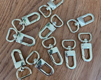 Puede incluir: Diez cierres de metal dorado giratorios, también conocidos como broches de presión o cierres de langosta, están dispuestos sobre una superficie de madera. Estos cierres se utilizan comúnmente para sujetar correas, cadenas u otros accesorios.