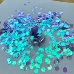 Könnte beinhalten: Ein kleines Glas mit schimmerndem blauem und violettem Glitter, verstreut auf einer hellgrauen Oberfläche. Der Glitter hat die Form kleiner Sechsecke.