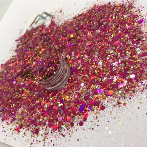 May include: A close-up of a jar of pink, gold, and iridescent glitter. The glitter is scattered on a white surface.