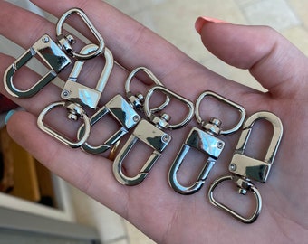 Puede incluir: Seis cierres de mosquetón de metal plateado se muestran en una mano. Los cierres se utilizan comúnmente para sujetar correas, cadenas u otros accesorios.