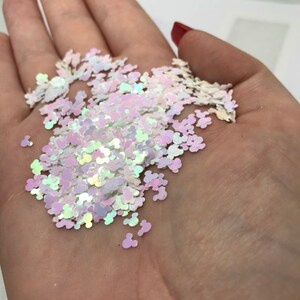 May include: A collection of small, iridescent, white, mouse-shaped confetti pieces. Each individual piece features a distinct three-circle silhouette, reflecting light with a pearlescent sheen that shifts between soft pink, blue, and yellow tones.