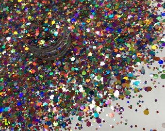 Op de afbeelding: Een close-up van een hoop iriserende glitter in verschillende vormen en maten, waaronder zeshoeken. De glitter is een mix van kleuren, waaronder goud, groen, blauw en paars. Een kleine, doorzichtige pot is gedeeltelijk zichtbaar, gevuld met de glitter.