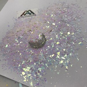 May include: A close-up of iridescent, white, and clear chunky glitter. The glitter is scattered on a white surface.