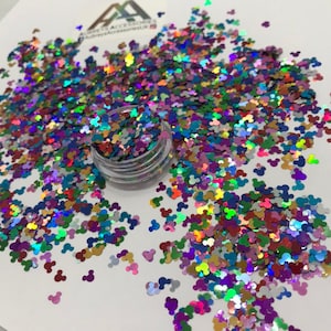 Puede incluir: Un pequeño frasco de purpurina multicolor iridiscente con forma de cabezas de Mickey Mouse. La purpurina está esparcida sobre una superficie blanca.