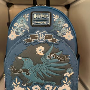 May include: A blue mini backpack featuring a Ravenclaw eagle emblem, floral embroidery, and banners with the words WIT, WISDOM, and LEARNING. The bag includes a Harry Potter Loungefly metal logo plate, a top handle, and silver-toned hardware accents.