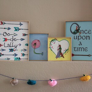 May include: Four framed pictures on a wall with a garland of yarn balls hanging below. The pictures are all different sizes and have different designs. The first picture has a white background with colourful arrows and the text "Oo-de-Lally". The second picture has a blue background with a pink flower and a green stem. The third picture has a yellow background with a heart shape and a couple inside. The fourth picture has a blue background with the text "Once upon a time" and a single arrow.