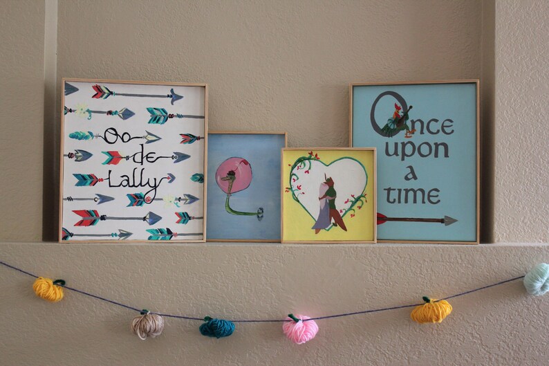 May include: Four framed pictures on a wall with a garland of yarn balls hanging below. The pictures are all different sizes and have different designs. The first picture has a white background with colourful arrows and the text "Oo-de-Lally". The second picture has a blue background with a pink flower and a green stem. The third picture has a yellow background with a heart shape and a couple inside. The fourth picture has a blue background with the text "Once upon a time" and a single arrow.