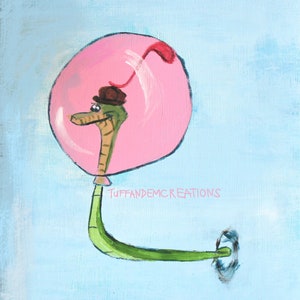May include: A green snake wearing a brown hat floats in a pink bubble with a red tail. The snake is facing left and has a small, black eye. The background is a light blue colour. The text "TUFFANDEMCREATIONS" is written in black below the bubble.