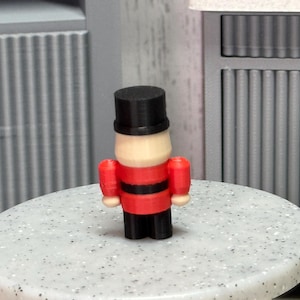 May include: A small, toy soldier figurine with a black top hat, a red jacket with black stripes, and black pants. The toy soldier is standing on a speckled, light-colored surface. The background features a gray cabinet.