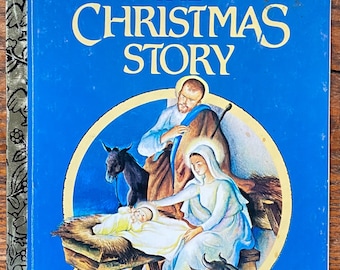 May include: A Little Golden Book titled "The Christmas Story" with a blue cover and an illustration of the nativity scene. The illustration shows Mary, Joseph, and the baby Jesus in a manger with a donkey and an ox.