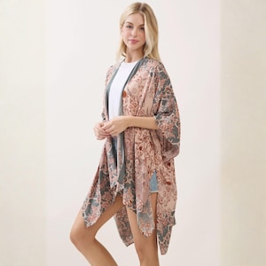 May include: A lightweight open-front kimono featuring a muted sage green and dusty rose floral damask pattern. The garment has wide, elbow-length sleeves and a relaxed, flowing silhouette that drapes over a plain white t-shirt and denim shorts.