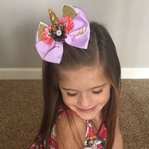 May include: A lavender unicorn-themed hair bow with a gold horn and accents. The bow features gold glitter accents, a central floral arrangement, and is worn in the hair. The bow is a whimsical accessory.