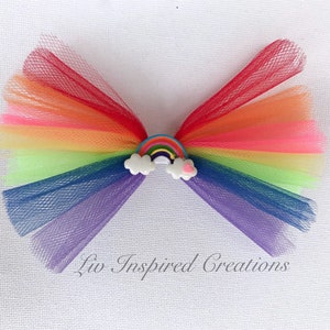 Può includere: Un fiocco per capelli a tema arcobaleno realizzato in tulle a strati rossi, arancioni, gialli, verdi, blu e viola. Il fiocco presenta un piccolo arcobaleno e un ornamento a nuvola al centro. Il testo "Liv Inspired Creations" è stampato in basso.