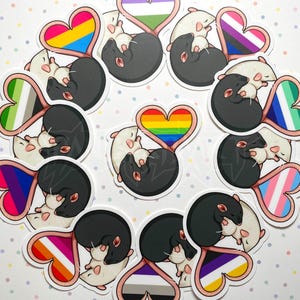 May include: A collection of stickers featuring cartoon rats in a heart shape. Each rat is black and white, with a heart-shaped tail in various pride flag colors, including rainbow, transgender, and bisexual flags. The stickers are arranged in a circular pattern.
