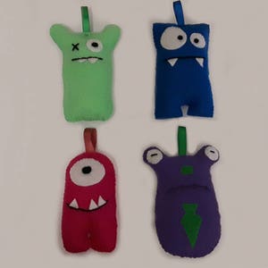 May include: Four felt monster ornaments with different colors and facial expressions. The monsters are green, blue, red, and purple.