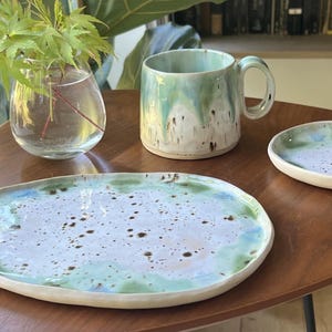 Puede incluir: Un juego de cerámica compuesto por una taza, un plato ovalado grande y un plato redondo pequeño. Cada pieza presenta una base blanca con un esmalte reactivo en tonos verde claro y azul, acentuado por motas de color marrón oscuro.
