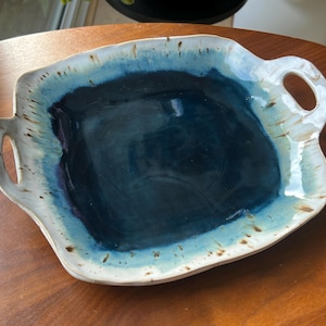 May include: A square ceramic serving tray with two integrated handles. The centre features a deep navy blue glaze that transitions into a lighter turquoise and white rim, accented with brown speckles along the wavy, uneven edges of the piece.