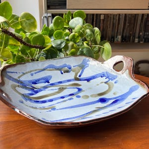 May include: A shallow, hand-formed ceramic serving tray with a light grey speckled base. The surface features abstract, flowing brushstrokes in deep blue and muted olive green. It has a dark brown, uneven rim and two integrated side handles.