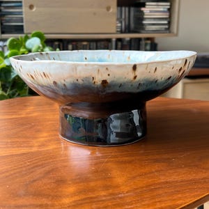 May include: A ceramic pedestal bowl featuring a wide, shallow basin with an irregular, wavy rim. The exterior and interior glaze transitions from a deep, dark brown base to a mottled blue and white upper section with brown speckles throughout.