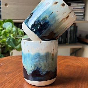 May include: Two cylindrical ceramic cups stacked vertically. The cups feature a reactive glaze with a colour palette of deep navy blue, teal, and cream white. The glaze drips down the sides, creating a layered effect over the smooth clay base.