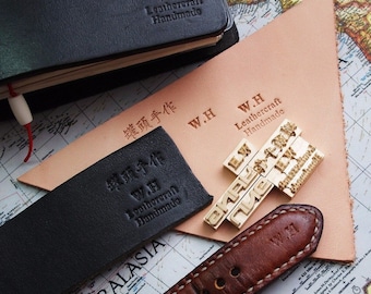 May include: Close-up of a leather craft stamp set with the text "W.H Leathercraft Handmade" in gold lettering. The stamps are arranged on a light brown leather surface. The image also shows a black leather journal with the same text stamped on the cover.