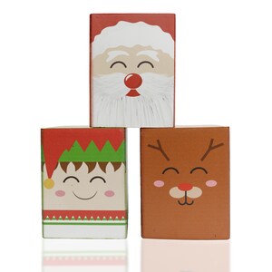 May include: Three wooden blocks stacked on top of each other. The top block features a red and white Santa Claus face. The middle block features a green elf face with a red hat. The bottom block features a brown reindeer face with antlers.