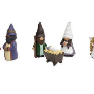 May include: A collection of ceramic Christmas gnomes in various colors. The set includes gnomes dressed as the three wise men, an angel, a shepherd with a sheep, and a gnome with a baby in a manger. The figures have detailed features and are set against a white background.