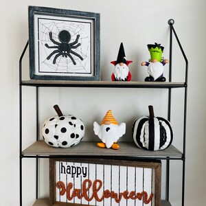 K&ouml;nnte beinhalten: Ein dreistufiges Regal mit Halloween-Dekorationen. Auf den Regalen befinden sich ein gerahmtes Spinnennetz-Bild, Zwergfiguren und Stoffk&uuml;rbisse. Ein Schild mit der Aufschrift "Happy Halloween" befindet sich unten.
