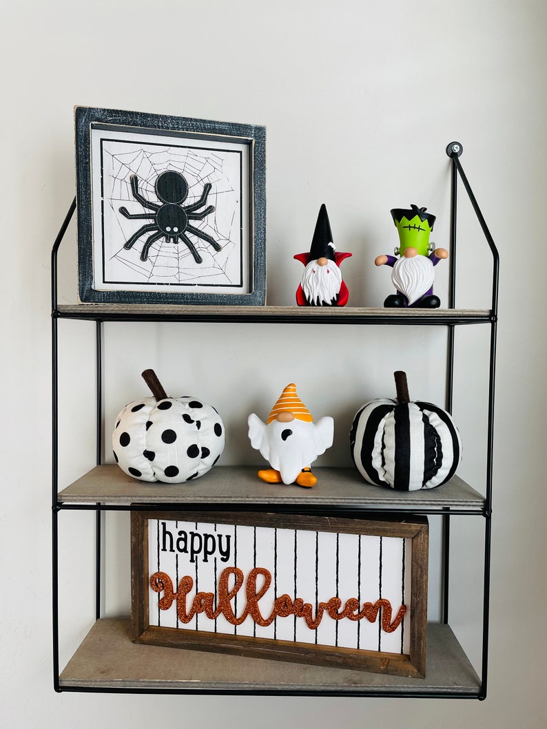 K&ouml;nnte beinhalten: Ein dreistufiges Regal mit Halloween-Dekorationen. Auf den Regalen befinden sich ein gerahmtes Spinnennetz-Bild, Zwergfiguren und Stoffk&uuml;rbisse. Ein Schild mit der Aufschrift "Happy Halloween" befindet sich unten.
