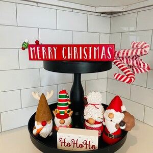 May include: A black tiered tray with Christmas decorations. The top tier has a red sign that says "MERRY CHRISTMAS" and candy canes. The bottom tier has gnomes and a sign that says "Ho Ho Ho".
