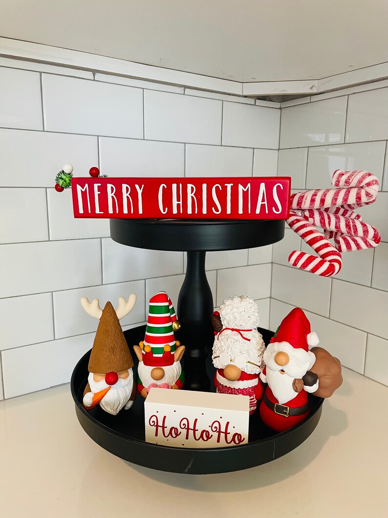 May include: A black tiered tray with Christmas decorations. The top tier has a red sign that says "MERRY CHRISTMAS" and candy canes. The bottom tier has gnomes and a sign that says "Ho Ho Ho".