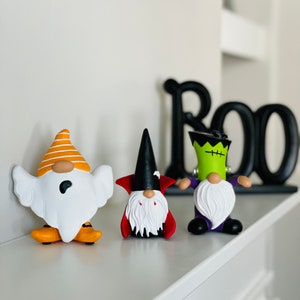K&ouml;nnte beinhalten: Drei Halloween-Zwergfiguren: ein Geist, ein Vampir und Frankensteins Monster. Der Geisterzwerg hat einen orange-wei&szlig; gestreiften Hut und orangefarbene Schuhe. Der Vampirzwerg hat einen schwarzen Hut und einen roten Umhang. Der Frankenstein-Zwerg hat gr&uuml;ne Haut und eine lila Jacke.
