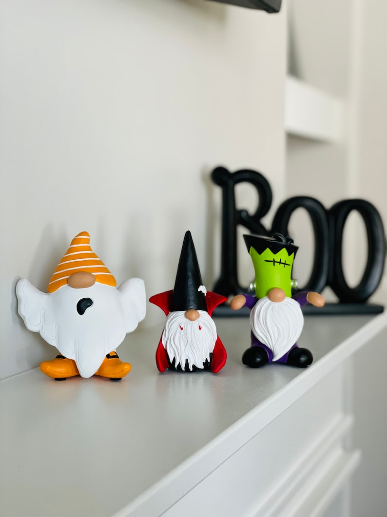 K&ouml;nnte beinhalten: Drei Halloween-Zwergfiguren: ein Geist, ein Vampir und Frankensteins Monster. Der Geisterzwerg hat einen orange-wei&szlig; gestreiften Hut und orangefarbene Schuhe. Der Vampirzwerg hat einen schwarzen Hut und einen roten Umhang. Der Frankenstein-Zwerg hat gr&uuml;ne Haut und eine lila Jacke.