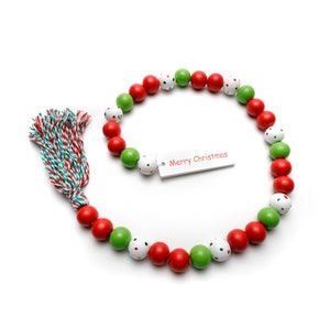 May include: A Christmas garland featuring red, green, and white wooden beads. Some beads have white polka dots. The garland includes a tassel and a tag that reads "Merry Christmas".