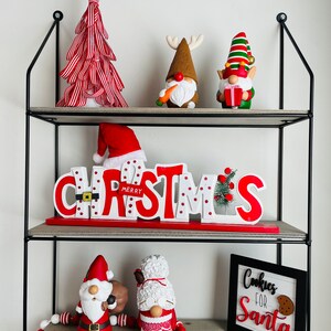 May include: A three-tiered shelf unit displays Christmas decorations. A red and white striped tree sits atop, with gnome figurines and a "Merry Christmas" sign below. A sign that says "Cookies for Santa" is on the bottom shelf.