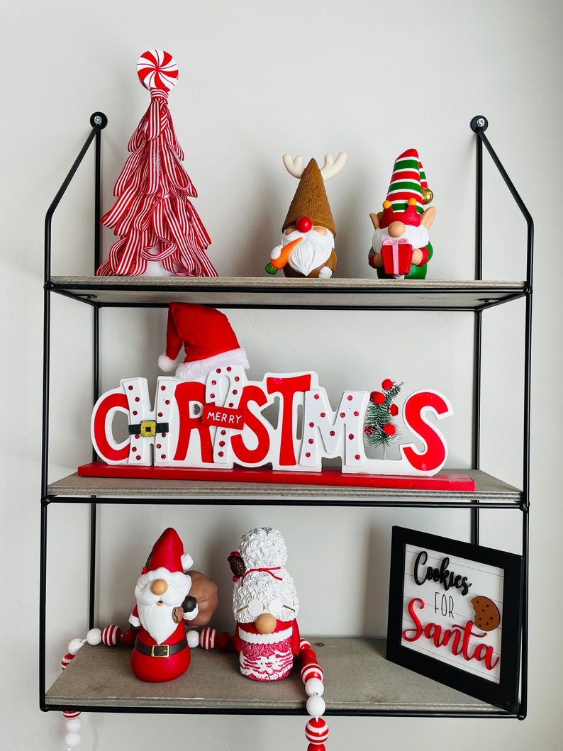 May include: A three-tiered shelf unit displays Christmas decorations. A red and white striped tree sits atop, with gnome figurines and a "Merry Christmas" sign below. A sign that says "Cookies for Santa" is on the bottom shelf.