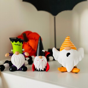 K&ouml;nnte beinhalten: Halloween-Themen-Zwergfiguren. Die Zwerge sind als Frankenstein, Vampir und Geist verkleidet. Der Frankenstein-Zwerg ist gr&uuml;n mit lila Akzenten, der Vampir-Zwerg hat einen schwarzen Umhang und der Geister-Zwerg ist wei&szlig; mit orangefarbenen Akzenten.