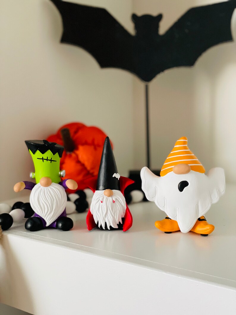 K&ouml;nnte beinhalten: Halloween-Themen-Zwergfiguren. Die Zwerge sind als Frankenstein, Vampir und Geist verkleidet. Der Frankenstein-Zwerg ist gr&uuml;n mit lila Akzenten, der Vampir-Zwerg hat einen schwarzen Umhang und der Geister-Zwerg ist wei&szlig; mit orangefarbenen Akzenten.