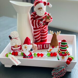 May include: A white tray holds Christmas decorations, including a Santa Claus figurine in a red and white striped suit, and several gnome figurines. A white pitcher is in the background. A tag reads "Merry Christmas."