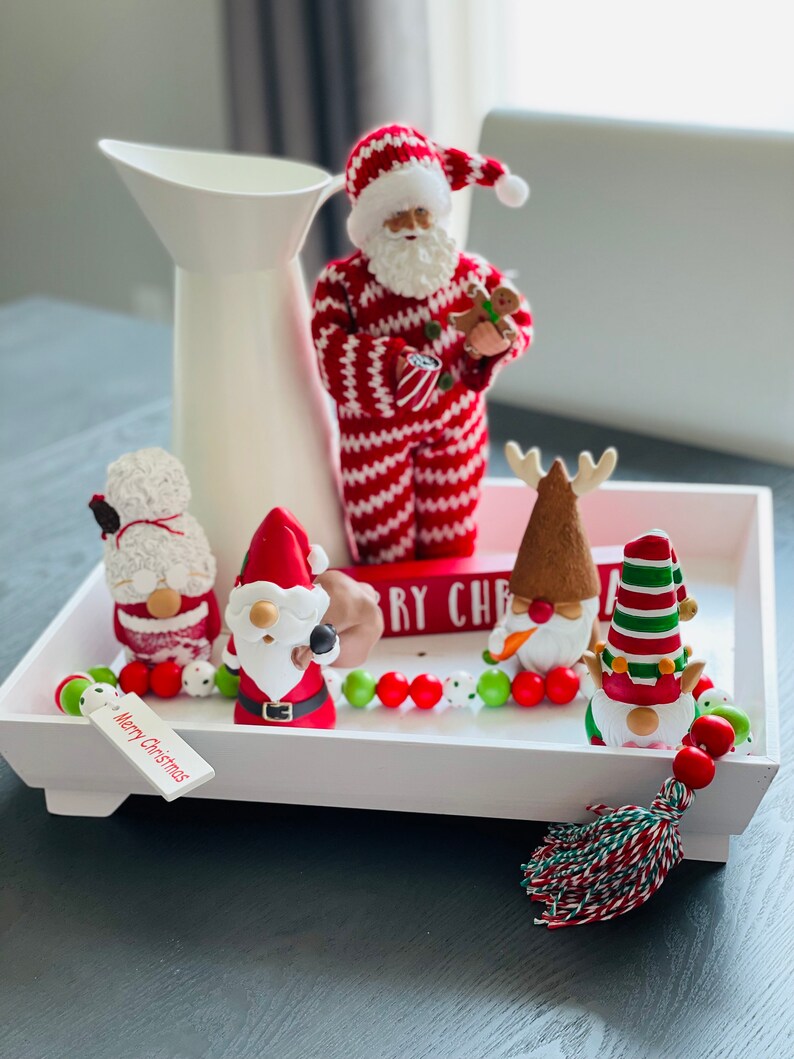 May include: A white tray holds Christmas decorations, including a Santa Claus figurine in a red and white striped suit, and several gnome figurines. A white pitcher is in the background. A tag reads "Merry Christmas."