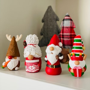 May include: A collection of Christmas gnome figurines with festive designs. The gnomes feature various outfits, including a reindeer, Santa Claus, and an elf, with details like a carrot, a gift, and a red and green striped hat. Behind them are fabric Christmas trees.