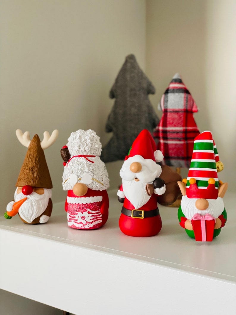 May include: A collection of Christmas gnome figurines with festive designs. The gnomes feature various outfits, including a reindeer, Santa Claus, and an elf, with details like a carrot, a gift, and a red and green striped hat. Behind them are fabric Christmas trees.