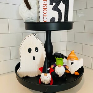 K&ouml;nnte beinhalten: Ein schwarzes, zweist&ouml;ckiges Tablett mit Halloween-Dekorationen. Die obere Ebene enth&auml;lt einen wei&szlig;en K&uuml;rbis und einen Blockkalender mit der Aufschrift "OKTOBER 31". Die untere Ebene zeigt einen Holzgeist und mehrere Zwergfiguren in Halloween-Kost&uuml;men.