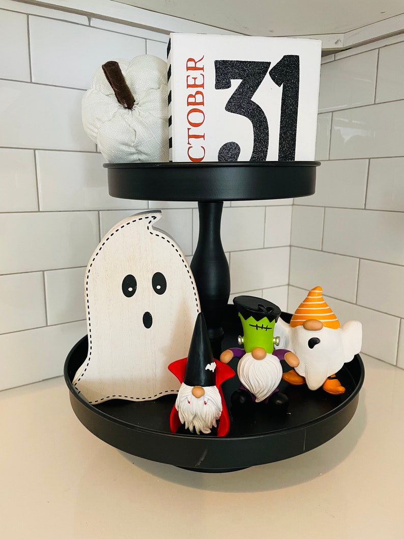 K&ouml;nnte beinhalten: Ein schwarzes, zweist&ouml;ckiges Tablett mit Halloween-Dekorationen. Die obere Ebene enth&auml;lt einen wei&szlig;en K&uuml;rbis und einen Blockkalender mit der Aufschrift "OKTOBER 31". Die untere Ebene zeigt einen Holzgeist und mehrere Zwergfiguren in Halloween-Kost&uuml;men.