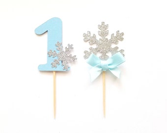 May include: Two blue and silver snowflake cake toppers. One topper features the number "1" with a small snowflake on the side. The other topper features a larger snowflake with a light blue bow.
