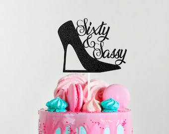 May include: A pink and blue cake with colorful meringue and macarons. A black glitter cake topper with a high heel shoe and the text "Sixty & Sassy" is on top of the cake.