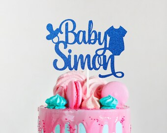 May include: Blue glitter cake topper with the text "Baby Simon" and a baby rattle and onesie design. The topper is on top of a pink and blue cake decorated with macarons and meringue cookies.