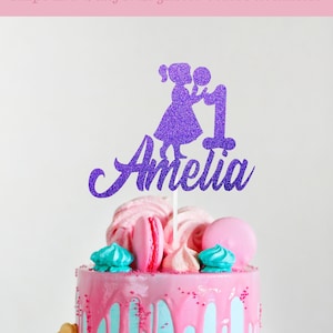 May include: Purple glitter cake topper with a girl holding a number one and the name Amelia. The topper is on a pink and blue cake with colorful decorations.