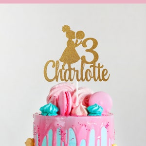 May include: A birthday cake with pink frosting dripping over a light blue base. The cake is decorated with macarons, meringues, and a gold glitter cake topper that reads "3 Charlotte". The cake sits on a white cake stand.