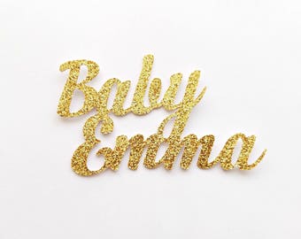 Może przedstawiać: Złote literki z brokatem, które tworzą napis "Baby Emma" na białym tle.